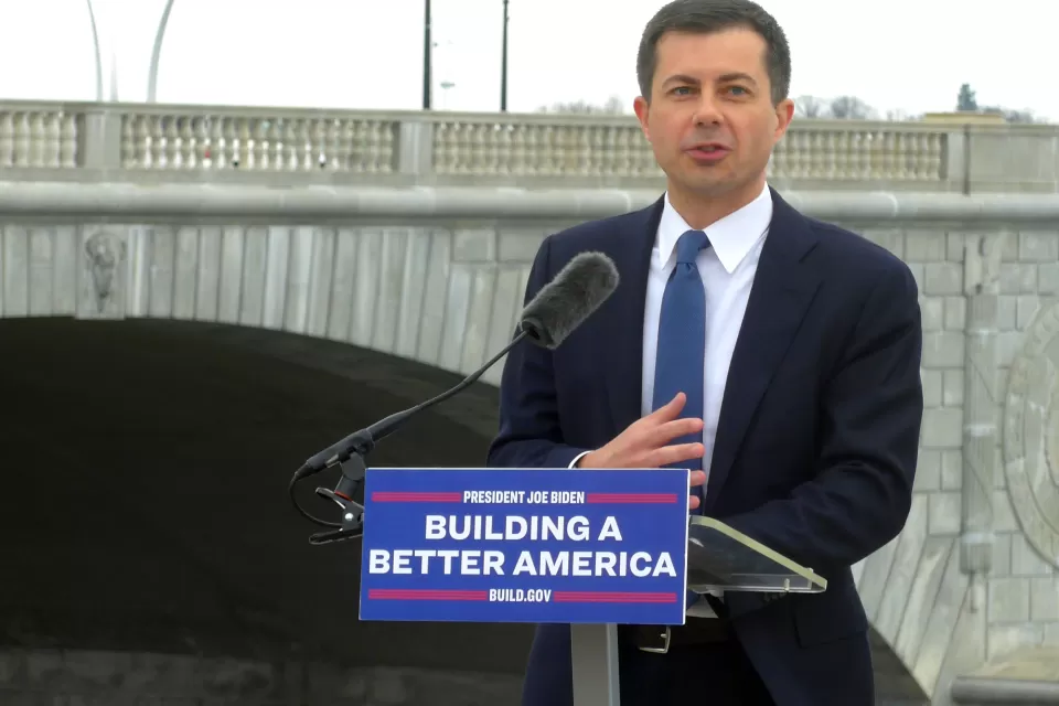 U.S> Department of Transportation Secretary Pete Buttigieg delivers a speech in front of a bridge in Washington, D.C. The podium where he stands has a sign that reads: "Building a Better America."